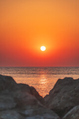 An orange sunset over the sea, the sun low on the horizon. A coastal landscape visible from the shore between the rocks.