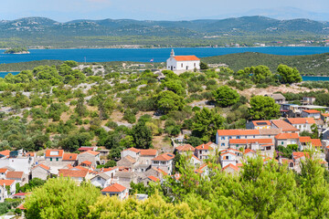 Church of St. Roch on a hill in the Croatian town of Murter, viewed from the top of Mount Raduc with mountains in the background on a sunny summer day.