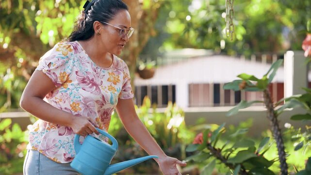 Smiling middle-aged woman enjoying her hobby, watering plants with a watering can in her lush green backyard during a sunny summer day