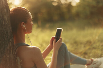 A young woman enjoys music on her smartphone while sitting outdoors under a tree, dressed in casual sporty attire, conveying relaxation and tranquility