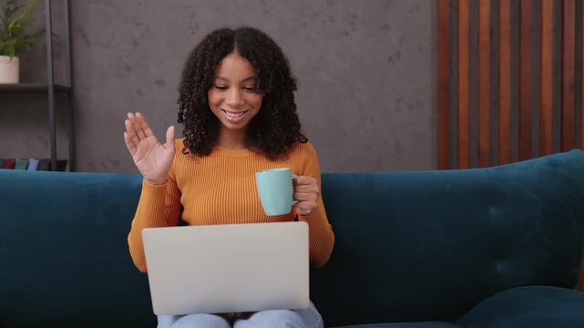 Little IT kid teen girl in casual clothes hold use work on laptop pc computer drink coffee watch movie sits on blue sofa couch stay at home hotel flat rest relax spend free spare time in living room