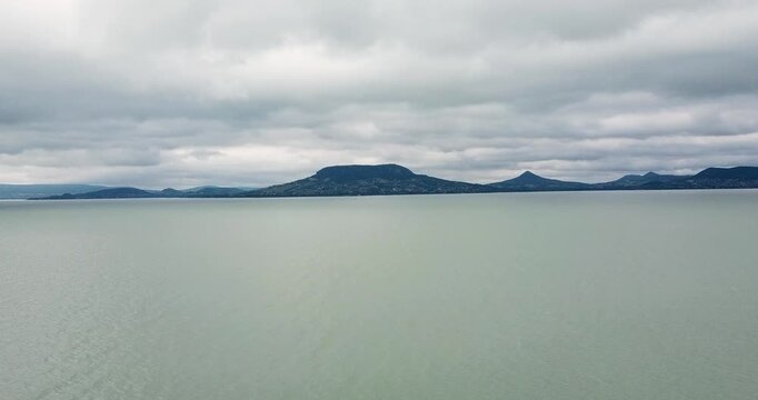 Aerial view of a calm Lake Balaton with a backdrop of distant hills and a cloudy sky, creating a serene yet melancholic atmosphere, Fonyod, Hungary.
