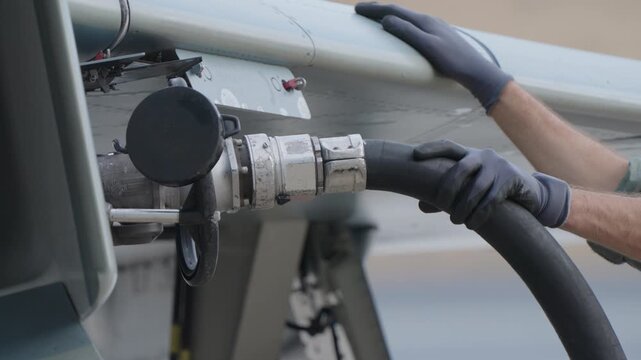 Military technician holding service hose connected to fighter jet to supply fuel or hydraulic fluid during controlled ground servicing procedure