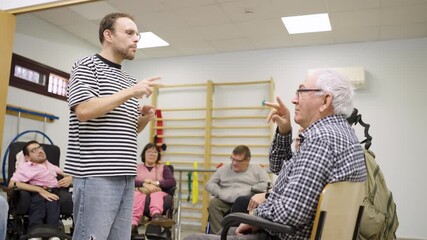Male social worker talking with a senior man in a wheelchair during a group therapy meeting in a support center for people with disabilities