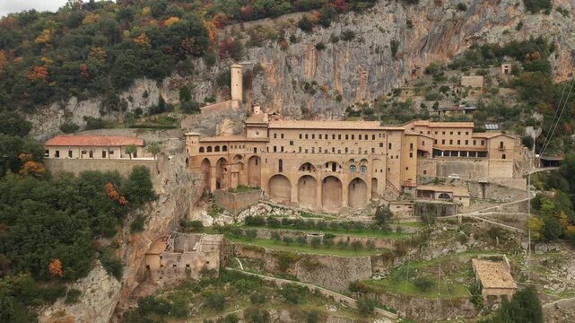 Aerial view of the Monastery of St. Benedict clinging to the rocky cliffside, blending with the autumn colors of the forest, Subiaco, Lazio, Italy.