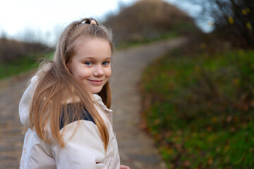 Portrait of a cheerful young girl walking on a path in nature, wearing a cream jacket, with greenery and soft natural light.