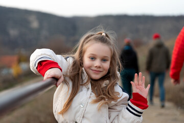 Cheerful young girl in a light jacket waving at the camera, standing by a railing with a blurred natural background, group of people in the distance.
