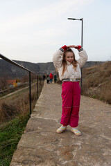Cheerful young girl in warm clothing poses on a stone path with hills in the background, evoking joy and adventure.