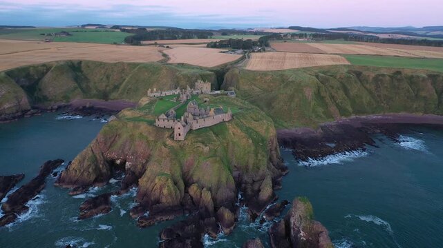Aerial view of Dunnottar Castle perched on a dramatic, rocky headland as waves crash against the cliffs, Stonehaven, Scotland, United Kingdom.
