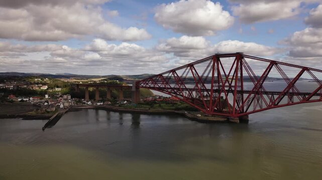 Aerial view of the striking red Forth Bridge spanning the Firth of Forth, contrasting with the green shoreline and cloudy sky, Inverkeithing, Scotland, United Kingdom.