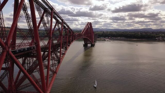 Aerial view of the majestic red Forth Bridge spanning the calm waters, with a sailboat gliding beneath, contrasting with cloudy skies, Inverkeithing, Scotland, United Kingdom.