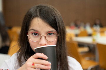 Schoolgirl in glasses drinking from paper cup while sitting in classroom, childhood education concept