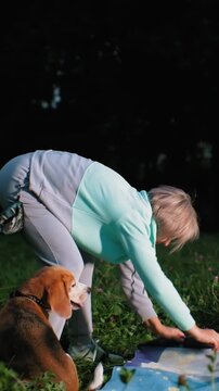 senior woman arranges illustrated blanket under dusk sky beagle sits close, woman smooths corners then settles to sit, soft distant lights, contemplative mood, gentle petting and shared quiet