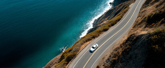 Aerial view of a white car driving along a winding coastal road with blue ocean waves crashing against the rocky shoreline
