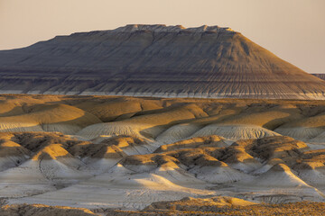 View of sun-kissed, undulating terrain with stark shadows and layered mesas under a soft sky paints a breathtaking scene, Senek, Mangystau Region, Kazakhstan.