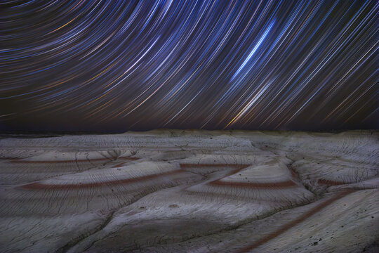 View of celestial star trails painting the night sky above the stark, layered landscape with red bands cutting through the arid ground, Senek, Mangystau Region, Kazakhstan.