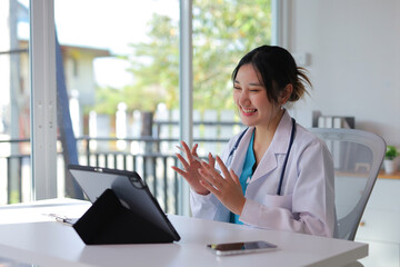 Smiling Asian female doctor wearing a lab coat and stethoscope is having a virtual consultation...