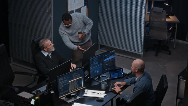 Three multiethnic cybersecurity workers interacting at desk in dark office with multiple computer screens displaying digital data and monitoring network security