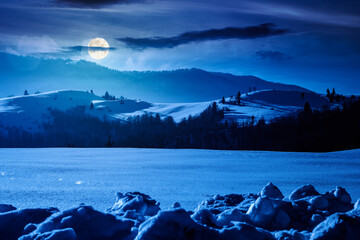 beautiful winter landscape with snow covered rolling hills under dark sky at night. majestic mountains in full moon light. frosty countryside scenery. backdrop for fiction art or mystery concepts