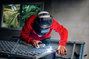 Welder wearing a red long-sleeved shirt and a protective mask is welding a steel grating, Industrial Welder With Torch