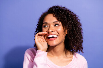 Young woman with curly hair wearing a pink sweater smiles brightly against a purple background for...