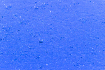 Blue surface covered with raindrops and circular ripples, single splash crowns frozen in falling rain.