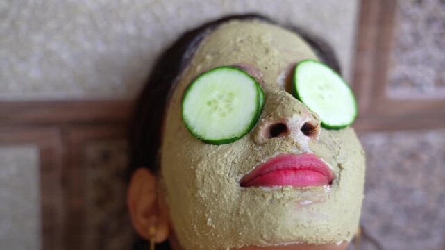A beautiful woman with facial clay mask on face and fresh cucumber on eyes relaxing at Home. Portrait of beautiful spa girl. Beauty, healthcare. Multani Mitti and green Face Pack. Asian Indian lady