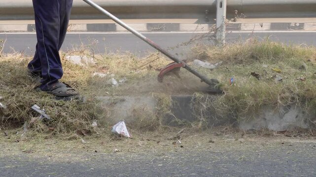person using a brush cutter, string trimmer or weed whacker to cut overgrown grass and weeds near a roadside. 