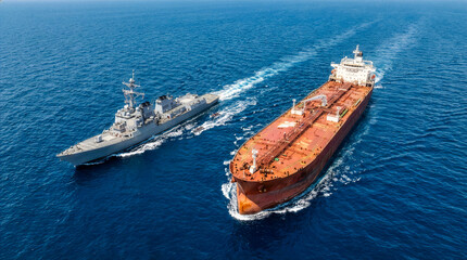 Military destroyer warship escorting large orange oil tanker vessel through blue ocean waters aerial view. Naval blockade maritime security operation concept.