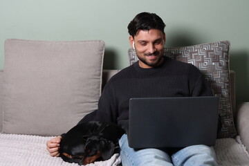 young man laying on sofa while working on laptop and petting his dog, remote work, work from home