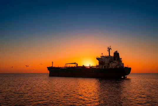 Oil tanker ship silhouette at sunset on open ocean. Maritime shipping cargo vessel at golden hour. International trade and naval blockade concept with dramatic sky.