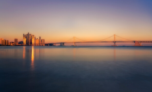 View of a long suspension bridge connecting to modern skyscrapers reflecting gold light in the calm sea at sunset, Busan, Busan, South Korea.
