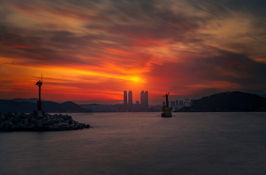 View of the fiery sunset casting a warm glow over the tranquil waters, silhouetting the city skyline and harbor structures, Busan, Busan, South Korea.