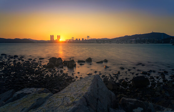 View of twilight paints the sky with fire above the tranquil waters and distant skyline, casting a warm glow on the rocky shore, Busan, Busan, South Korea.