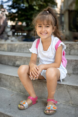 Adorable smiling little girl with a pink backpack sitting on the stairs outdoors. Happy first day of school or summer vacation portrait. Cheerful childhood moments.