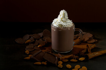 Hot chocolate cup with whipped cream surrounded by chocolate pieces and nuts on a dark surface during evening time