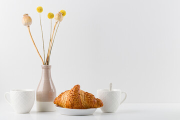 Croissant and a mug on a wooden stand, a vase , white background. minimalism. Copy space,