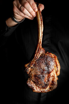 A tomahawk steak held by a person in dark attire, professionally grilled with a long bone, against a black background. This high-quality, close-up photo is perfect for food and dining themes