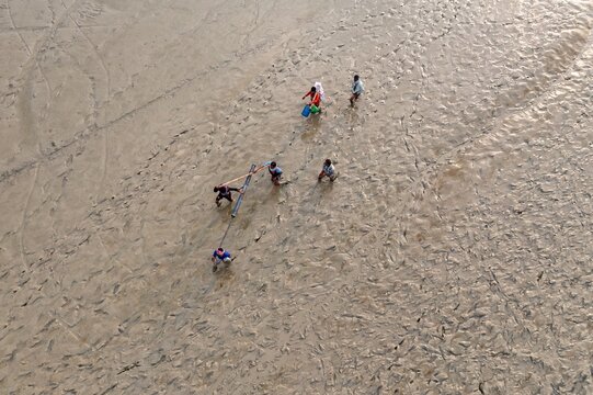Aerial view of a group of people scattered across the expansive muddy flats, their figures casting long shadows in the soft light, Chattogram, Chittagong Division, Bangladesh.