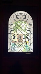 Ornate arched window with decorative wrought iron grille, seen from inside a dark interior. Elegant architectural detail with vintage design and soft daylight outside.