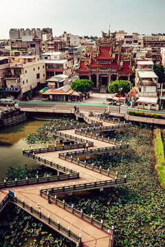 View of a zig-zag bridge cuts through a lily pad-covered waterway leading to a traditional temple in a bustling urban setting, Taipei City, Taiwan.