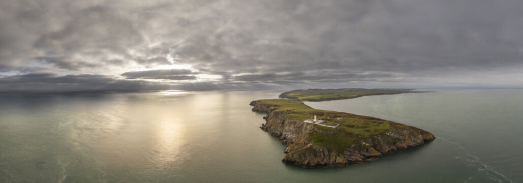Aerial view of the Mull of Galloway Lighthouse standing sentinel on a rugged, grassy headland as the sun pierces through a dramatic sky, Stranraer, Scotland, United Kingdom.
