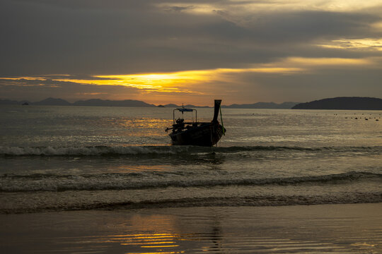 View of a longtail boat silhouette sits serenely on the water as the sun sets, casting a golden glow over the undulating waves, Ao Nang, Krabi, Thailand.