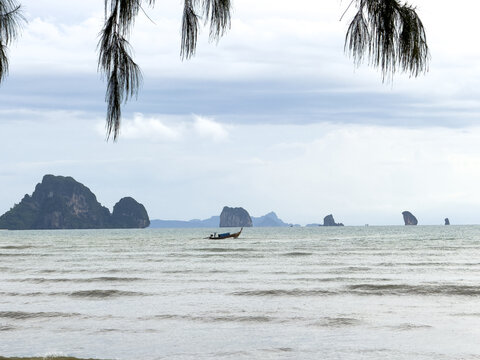 View of the tranquil sea reflecting a muted sky, adorned with distant islands and a traditional longtail boat, framed by overhanging foliage, Ao Nang, Krabi, Thailand.