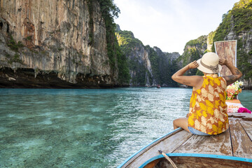 View of a woman in a yellow top and hat sits on the front of a wooden boat sailing through turquoise waters lined with towering cliffs, Phi Phi Island, Krabi, Thailand.