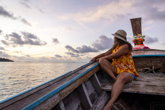 View of a woman with a hat and yellow dress sits on a wooden boat as the sun sets over the calm sea, painting the sky in soft hues, Phi Phi Island, Krabi, Thailand.