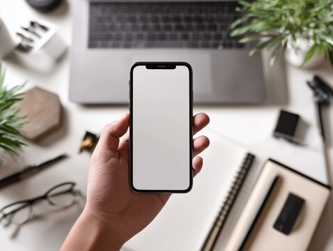Top angle hand holding smartphone with blank screen on office desk with laptop and stationery.