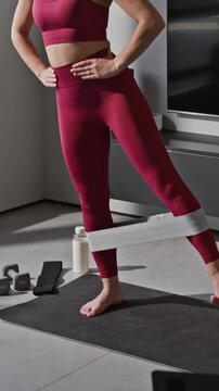 Vertical zoom in shot of Caucasian athletic woman doing single leg hip abduction using fitness band while exercising at home