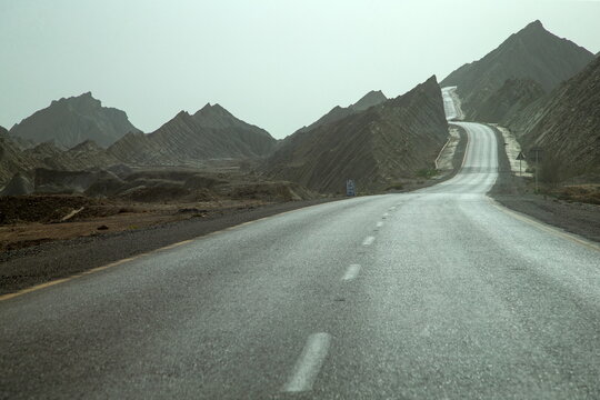 View of sun-kissed asphalt winds through rugged, arid mountains under a hazy sky, promising adventure on the Makran Coastal Highway, Balochistan, Pakistan.