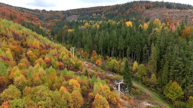 Aerial view of ski lift moving over autumn forest in mountains, colorful fall trees on hills landscape under cloudy sky at morning